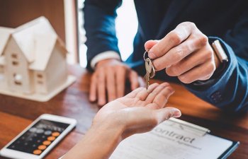 real estate agent holding with keys in his hand sitting at a desk with a house model on it